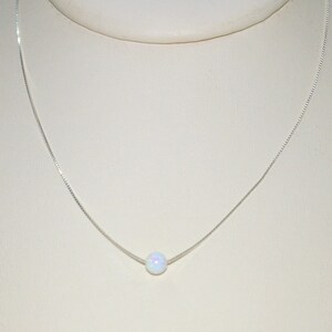 May include: A delicate silver necklace featuring a single, round, iridescent opal pendant. The opal has a milky white base with subtle flashes of color. The chain is a fine, box-style design, creating a minimalist and elegant aesthetic.