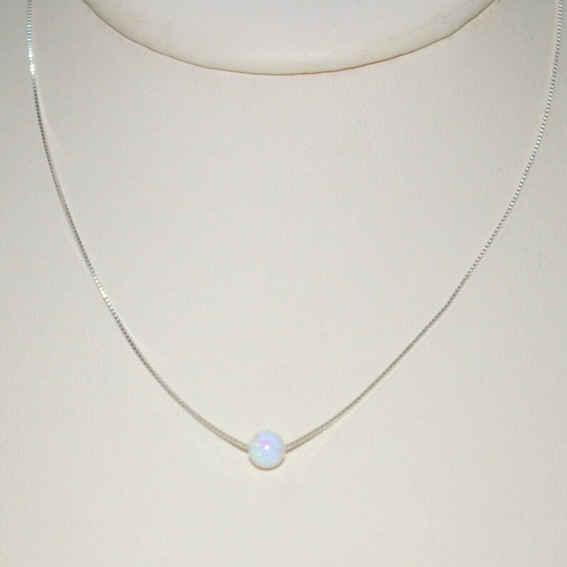 May include: A delicate silver necklace featuring a single, round, iridescent opal pendant. The opal has a milky white base with subtle flashes of color. The chain is a fine, box-style design, creating a minimalist and elegant aesthetic.