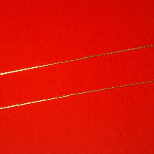 Puede incluir: Dos cadenas de oro sobre un fondo rojo. Las cadenas son finas y delicadas, con un diseño trenzado.