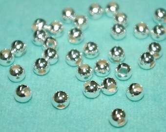 Könnte beinhalten: Eine große Menge silberfarbener Metallperlen, verstreut auf einer türkisfarbenen Oberfläche. Die Perlen sind rund und haben ein kleines Loch in der Mitte.