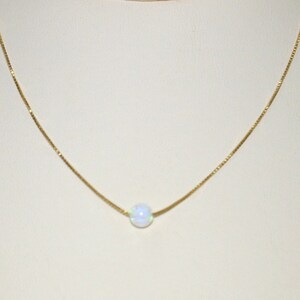 May include: A delicate gold necklace featuring a single, round, iridescent opal pendant. The opal has a soft, milky white color with subtle flashes of blue and green. The chain is a fine, gold box chain.