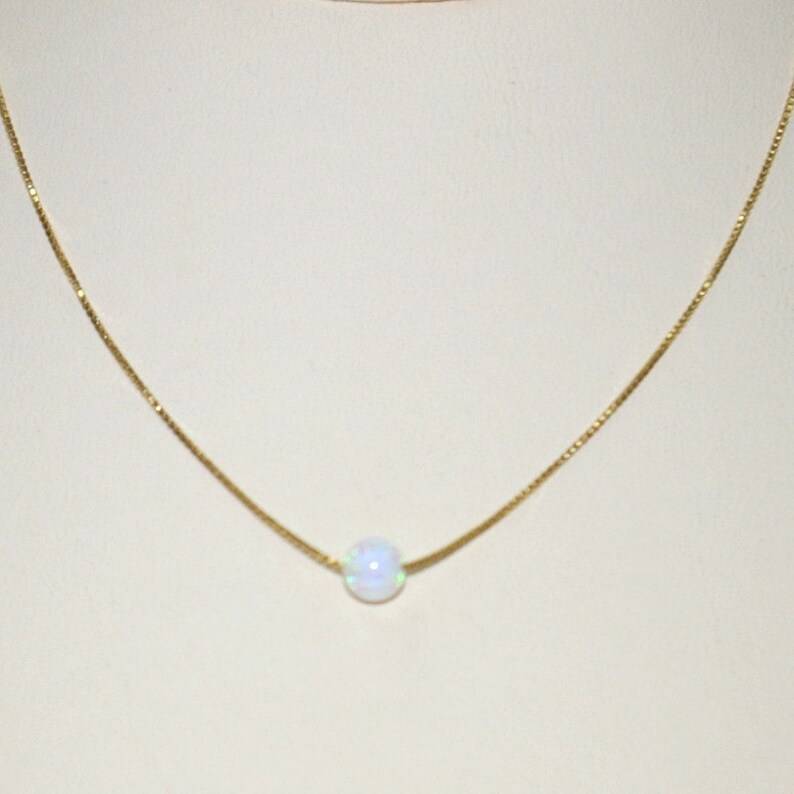May include: A delicate gold necklace featuring a single, round, iridescent opal pendant. The opal has a soft, milky white color with subtle flashes of blue and green. The chain is a fine, gold box chain.