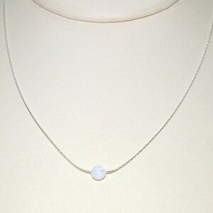 May include: A delicate silver necklace featuring a single, round, white opal pendant. The opal has subtle iridescent colors. The chain is thin and appears to be made of silver. The necklace is displayed against a neutral background.