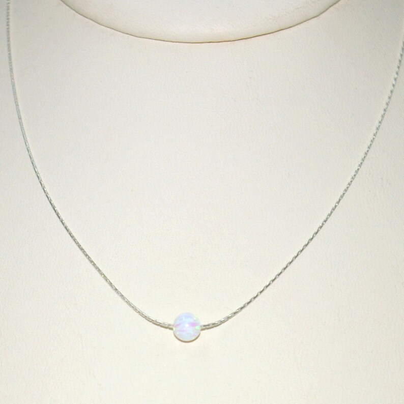 May include: A delicate silver necklace featuring a single, round, white opal pendant. The opal has subtle iridescent colors. The chain is thin and appears to be made of silver. The necklace is displayed against a neutral background.