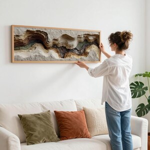 May include: A woman hangs a framed, layered artwork on a white wall. The artwork features a textured, abstract landscape in shades of brown, beige, and gray. The frame is made of light-colored wood. A white sofa with colorful pillows sits below the artwork.