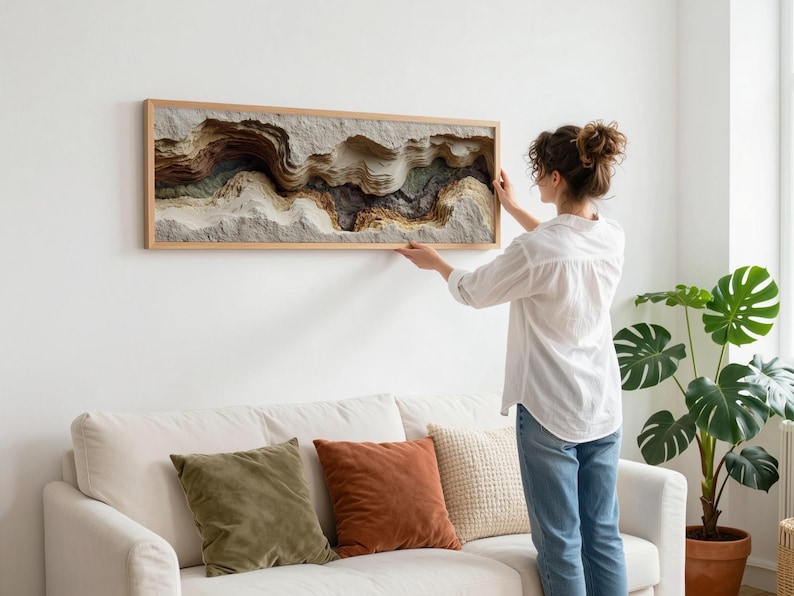 May include: A woman hangs a framed, layered artwork on a white wall. The artwork features a textured, abstract landscape in shades of brown, beige, and gray. The frame is made of light-colored wood. A white sofa with colorful pillows sits below the artwork.