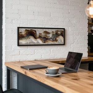 May include: A framed abstract art piece with a black frame hangs on a white brick wall. The artwork features a textured, layered design in shades of brown, beige, and green. A laptop, tablet, and coffee cup sit on a wooden counter.