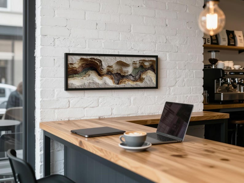 May include: A framed abstract art piece with a black frame hangs on a white brick wall. The artwork features a textured, layered design in shades of brown, beige, and green. A laptop, tablet, and coffee cup sit on a wooden counter.