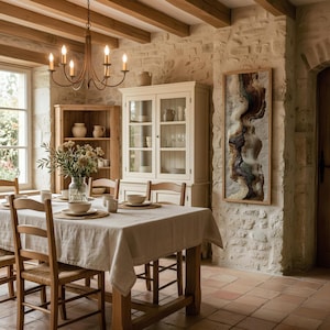 May include: A dining room scene with a rustic aesthetic. A wooden table is set with place settings and a linen tablecloth. Wooden chairs surround the table. A chandelier hangs above. A cabinet and artwork are visible against a stone wall.