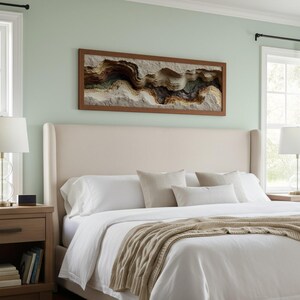 May include: A bedroom scene featuring a large bed with a cream-colored headboard and white bedding. A framed artwork with a textured, layered design hangs above the bed. Two bedside tables with lamps flank the bed, creating a cozy and inviting atmosphere.
