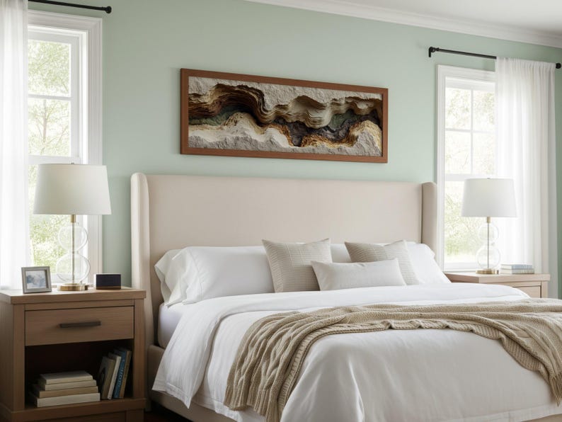 May include: A bedroom scene featuring a large bed with a cream-colored headboard and white bedding. A framed artwork with a textured, layered design hangs above the bed. Two bedside tables with lamps flank the bed, creating a cozy and inviting atmosphere.