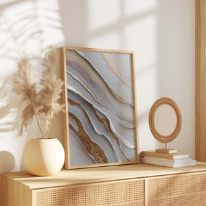 May include: A framed abstract art print with a textured surface. The artwork features flowing lines in shades of white, gray, gold, and purple. The frame is a light wood color, and the artwork is displayed on a wooden cabinet.