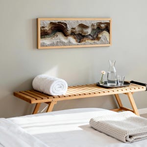 May include: A wooden bench with a rolled white towel, a tray with a water carafe and glasses, and a framed artwork depicting a layered landscape. A folded blanket rests on the bed in the foreground. The room has a neutral color palette.