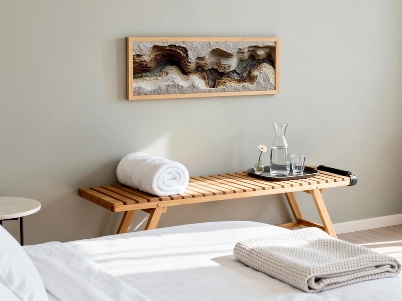 May include: A wooden bench with a rolled white towel, a tray with a water carafe and glasses, and a framed artwork depicting a layered landscape. A folded blanket rests on the bed in the foreground. The room has a neutral color palette.