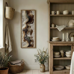 May include: A framed abstract art piece with a textured, layered design in shades of brown, beige, and green hangs on a light-colored wall. The artwork is rectangular and vertically oriented. A woven basket and a wooden cabinet are also visible.