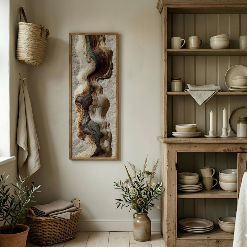 May include: A framed abstract art piece with a textured, layered design in shades of brown, beige, and green hangs on a light-colored wall. The artwork is rectangular and vertically oriented. A woven basket and a wooden cabinet are also visible.