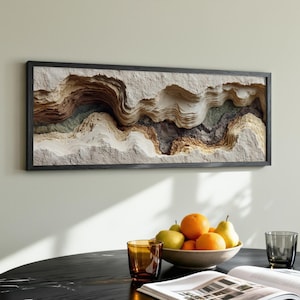 May include: A framed, abstract wall art piece with a layered, topographic design in shades of beige, brown, and gray. The artwork is mounted on a light-colored wall above a black table. A bowl of fruit and glasses are on the table.