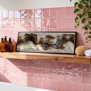 May include: A wooden shelf displays a framed abstract art piece, amber glass bottles, a natural sponge, and a rolled white towel. The shelf is set against a pink tiled wall, with a trailing green plant adding a touch of nature to the bathroom setting.
