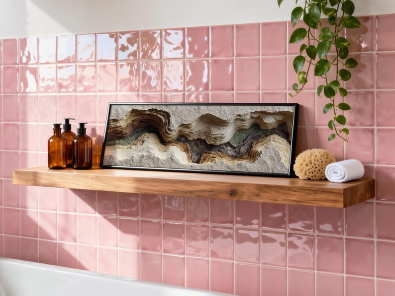 May include: A wooden shelf displays a framed abstract art piece, amber glass bottles, a natural sponge, and a rolled white towel. The shelf is set against a pink tiled wall, with a trailing green plant adding a touch of nature to the bathroom setting.