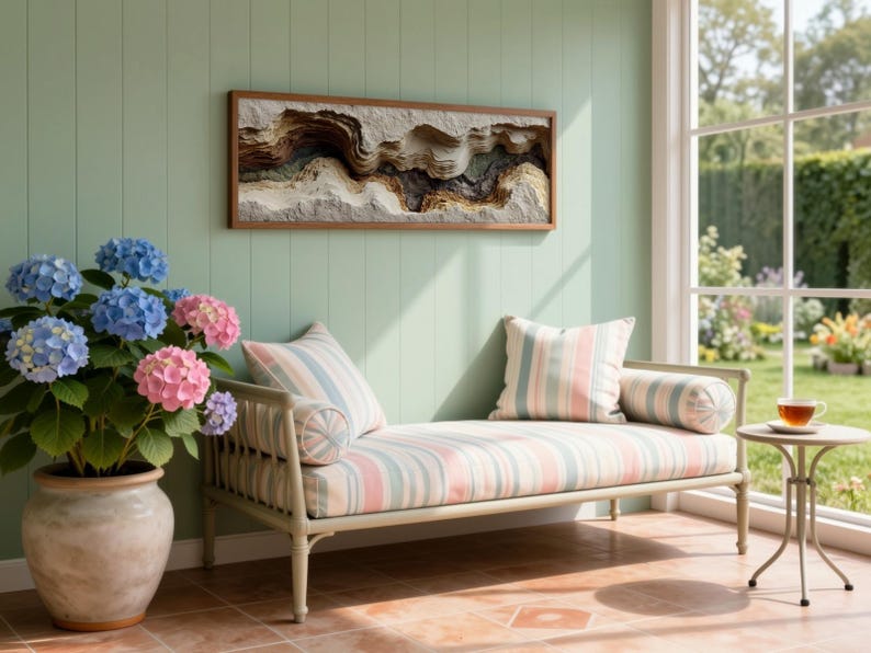 May include: A serene interior scene features a light green wall with a framed abstract art piece. A striped chaise lounge with pillows sits beneath the artwork. A potted hydrangea with blue and pink blooms adds a touch of color. A small side table holds a cup of tea.