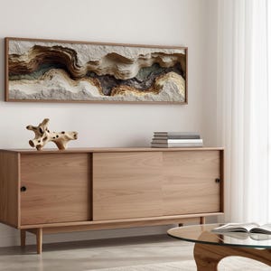 May include: A modern living room scene featuring a light wood sideboard with sliding doors, a round glass coffee table, and a framed abstract wall art piece. A decorative wooden sculpture and a stack of books are on the sideboard.