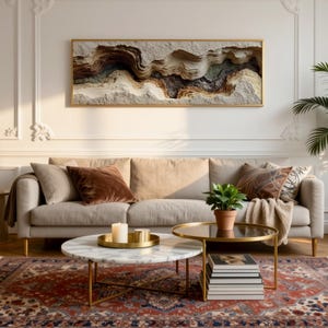 May include: A living room scene featuring a beige sofa with decorative pillows, a patterned rug, and a coffee table. A framed artwork with a textured, layered design hangs above the sofa. A potted plant and a side table complete the decor.
