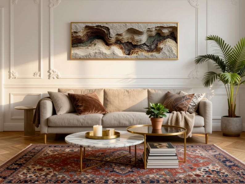 May include: A living room scene featuring a beige sofa with decorative pillows, a patterned rug, and a coffee table. A framed artwork with a textured, layered design hangs above the sofa. A potted plant and a side table complete the decor.