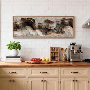 May include: A kitchen scene featuring a framed abstract art piece with layered textures in neutral tones. A white vase with a green plant sits on a stack of books. Fresh tomatoes and lemons are on a wooden cutting board. A spice rack and espresso machine are also visible.
