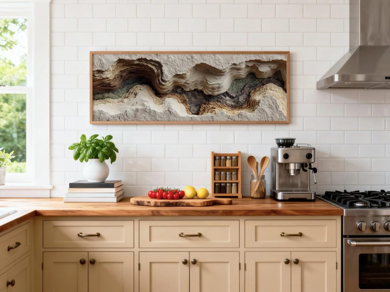 May include: A kitchen scene featuring a framed abstract art piece with layered textures in neutral tones. A white vase with a green plant sits on a stack of books. Fresh tomatoes and lemons are on a wooden cutting board. A spice rack and espresso machine are also visible.