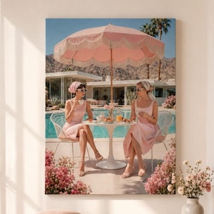 May include: A wall art print depicting two individuals in pink dresses and headscarves sitting at a white round table under a large pink fringed umbrella by a swimming pool, with a modern house and desert mountains in the background.