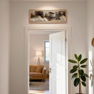 May include: A rectangular, framed artwork with a layered, abstract design in shades of brown, beige, and gray hangs above a doorway. A straw hat and scarf hang on a wall hook. A potted plant with large green leaves is in the foreground.