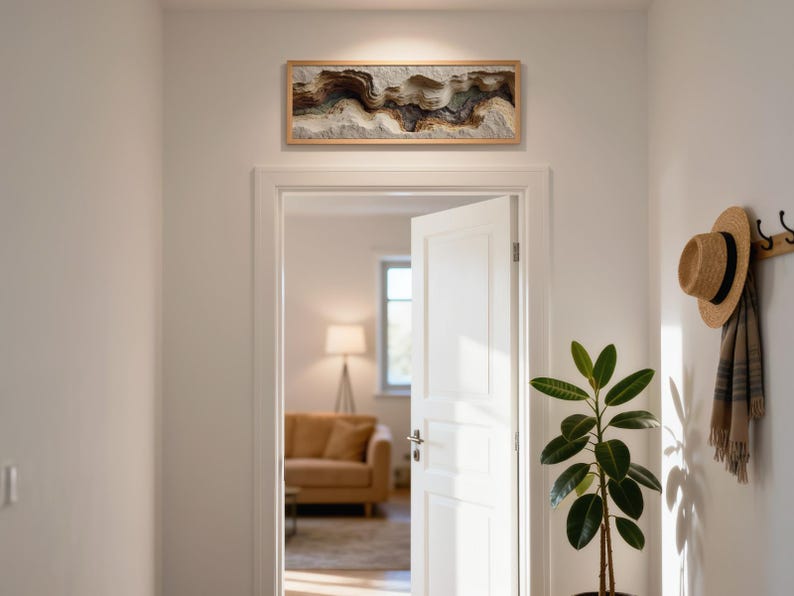May include: A rectangular, framed artwork with a layered, abstract design in shades of brown, beige, and gray hangs above a doorway. A straw hat and scarf hang on a wall hook. A potted plant with large green leaves is in the foreground.