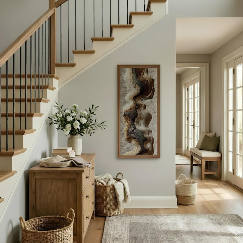 May include: A well-lit entryway with a wooden staircase, a framed abstract art piece, and a wooden chest of drawers. A white vase with flowers sits on the chest. Wicker baskets and a rug add to the decor.