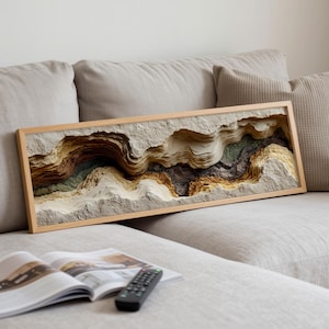 May include: A framed, layered paper art piece depicting a canyon landscape with shades of beige, brown, and green. The artwork is rectangular and rests on a light-colored sofa. A magazine and remote control are in the foreground.