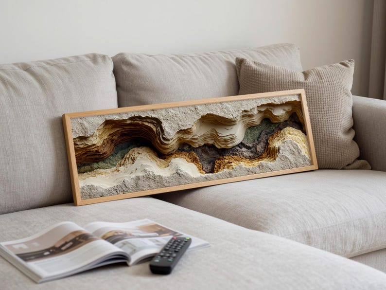 May include: A framed, layered paper art piece depicting a canyon landscape with shades of beige, brown, and green. The artwork is rectangular and rests on a light-colored sofa. A magazine and remote control are in the foreground.