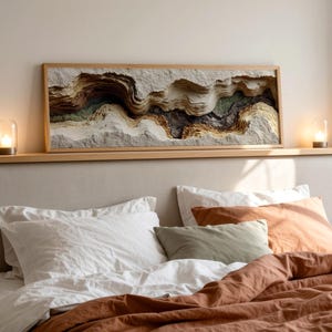 May include: A bedroom scene featuring a bed with white pillows, an orange pillow, and a rust-colored comforter. Above the bed is a wooden shelf with a framed, layered relief artwork in neutral tones. Two small glass lamps with lit candles sit on the shelf.