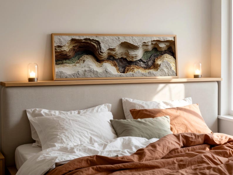 May include: A bedroom scene featuring a bed with white pillows, an orange pillow, and a rust-colored comforter. Above the bed is a wooden shelf with a framed, layered relief artwork in neutral tones. Two small glass lamps with lit candles sit on the shelf.
