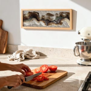 May include: A kitchen scene with a wooden cutting board and a person slicing tomatoes. A framed artwork with layered, textured design hangs on the wall. A cream-colored stand mixer is on the counter, along with a wooden cutting board.