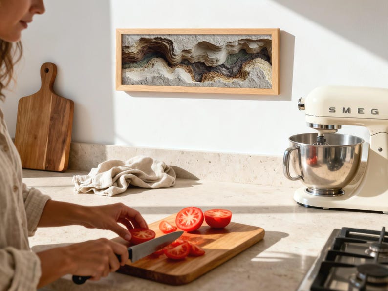 May include: A kitchen scene with a wooden cutting board and a person slicing tomatoes. A framed artwork with layered, textured design hangs on the wall. A cream-colored stand mixer is on the counter, along with a wooden cutting board.