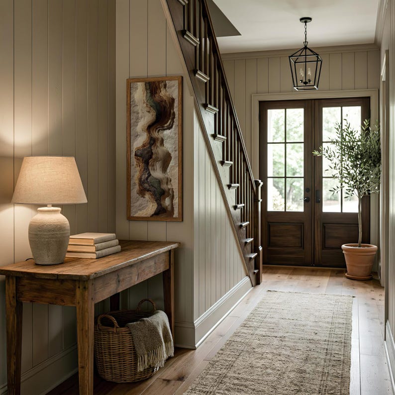May include: A hallway with a wooden staircase, a rustic wooden table with a lamp, books, and a basket. A framed abstract artwork hangs on the wall. The hallway has a neutral color palette with a rug and a potted plant.
