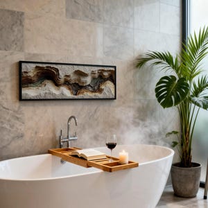 May include: A modern bathroom scene featuring a white oval bathtub with a wooden tray holding a book, a glass of red wine, and a lit candle. A framed artwork with a layered, textured design hangs above the tub. A potted plant and a towel rack complete the spa-like setting.