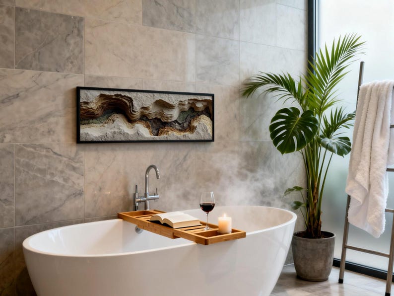 May include: A modern bathroom scene featuring a white oval bathtub with a wooden tray holding a book, a glass of red wine, and a lit candle. A framed artwork with a layered, textured design hangs above the tub. A potted plant and a towel rack complete the spa-like setting.
