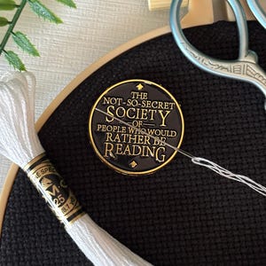 May include: A black embroidery hoop with white thread, scissors, and a pin that reads "The Not-So-Secret Society of People Who Would Rather Be Reading." The thread is labeled "M-C 25 18.7 yds."