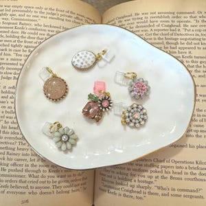 May include: A collection of decorative charms with various designs, including floral, crystal, and patterned elements. The charms are displayed on a white, organically shaped dish with a gold rim, set against the backdrop of an open book.
