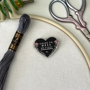 May include: A black heart-shaped enamel pin with the text "GOING TO HELL ON A FULL SCHOLARSHIP" and rose accents. The pin is on a white fabric background with embroidery supplies, including gray thread, scissors, and an embroidery hoop.