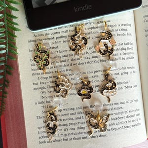 May include: A collection of gold-toned snake and butterfly charms with black and white enamel details. Some charms feature floral accents. The charms are displayed on clear acrylic stands, set against an open book and a Kindle.