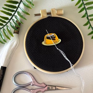 May include: A cross-stitch embroidery hoop with black fabric, featuring a yellow dessert pin. White thread and a needle are attached to the pin. A pair of pink and silver scissors and white embroidery floss are also present.