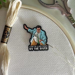 May include: An embroidery hoop with white fabric, cream-colored thread, and a needle. A pin with the text "I LIVE IN A VAN DOWN BY THE RIVER" is attached to the fabric. Scissors and other crafting supplies are also visible.