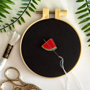 May include: A cross-stitch hoop with a black fabric, featuring a watermelon slice design. A needle with white thread is inserted into the fabric. Gold scissors and a spool of white thread are also present, along with green fern leaves.
