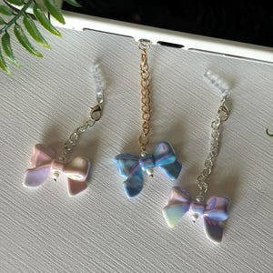 May include: Three iridescent bow charms with chains. The bows are in shades of pink, blue, and purple, each with a small pearl accent. The chains are silver and gold, designed to attach to a phone or other device.